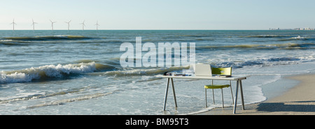 Schreibtisch und Stuhl mit Laptop am Strand, am Wasser setzen Windkraftanlagen am Horizont Stockfoto