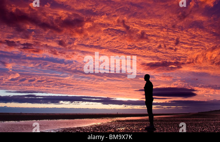 Antony Gormleys "Woanders" Crosby Strand Merseyside. Einzelzimmer "Iron Man" Silhouette gegen einen dramatischen roten Sonnenuntergang Stockfoto