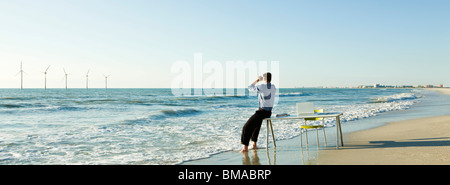 Mann sitzt am Rand des Schreibtisches am Rand des Wassers am Strand mit Blick auf Offshore-Windkraftanlagen am Horizont Stockfoto