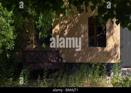 Altes Bauernhaus und Bank im Schatten eines Baumes in einem verwilderten Garten Stockfoto