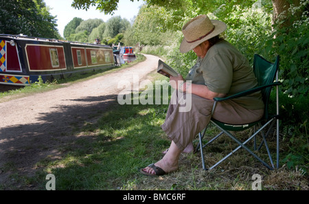 Sitzt eine Frau, canal entspannen und lesen ein Buch von Grand Union Leinpfad in der Nähe vor Anker schmale Boote Stockfoto