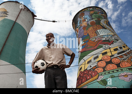 Ein Mann steht mit einem Fußball vor den Kühltürmen befindet sich in SOWETO, Johannesburg in Südafrika, Gauteng Stockfoto