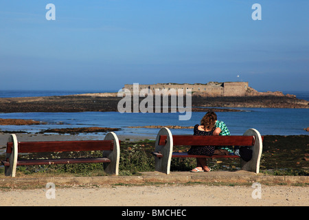 Junges Paar sitzt auf der Bank in Longis Bay mit Blick auf Ile de Raz Fort Alderney, Kanalinsel Vereinigtes Königreich Stockfoto