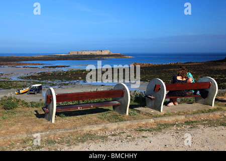 Junges Paar sitzt auf der Bank in Longis Bay mit Blick auf Ile de Raz Fort, Alderney, Kanalinsel Vereinigtes Königreich Stockfoto