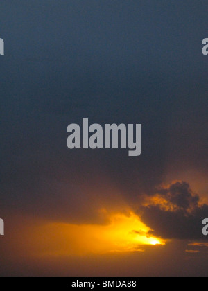 Die Sonne bricht durch, dass er Wolken bei Sonnenuntergang. Die Sonne ist hell Orange vor einem dunklen Himmel blau/grau. Stockfoto