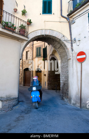 Mann reitet einen blaue Vespa Motorroller durch ein altes Tor in Spello, Provinz Perugia, Umbrien, Italien Stockfoto