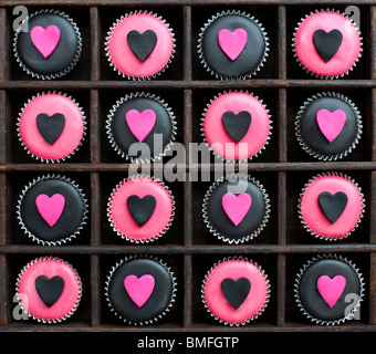 Mini-Muffins mit schwarz und rosa Zuckerguss und Herz Formen in einem Holztablett dekoriert Stockfoto