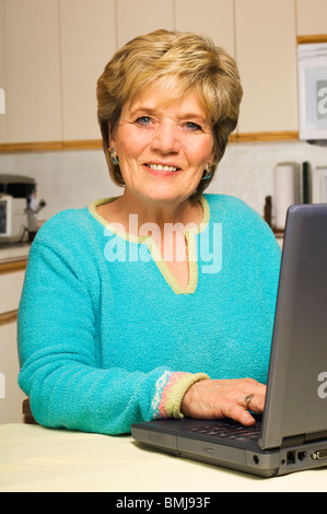 Eine ältere Frau lächelt während der Arbeit an ihrem Laptop in ihrer Küche Stockfoto