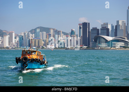 Ein Boot segelt in Richtung Betrachter im linken unteren Bereich des Rahmens mit einem Wald aus Wolkenkratzern als Kulisse in Hong Kong Stockfoto