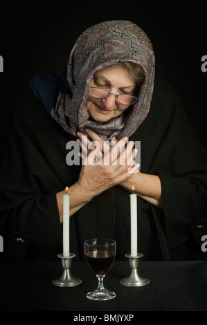 Eine ältere jüdische Frau, Kopf mit einem Tuch bedeckt, betet über zwei Sabbat-Kerzen und Sabbat Wein Stockfoto