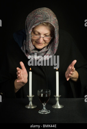 Eine ältere jüdische Frau, Kopf mit einem Tuch bedeckt, betet über zwei Sabbat-Kerzen und Sabbat Wein Stockfoto