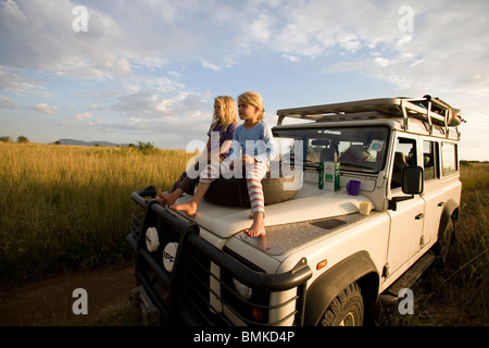 Afrika, Kenia, Masai Mara. Zwillingsmädchen sitzen auf der Motorhaube von ihrem Land Rover, wie sie den Blick über den Wildpark. Stockfoto