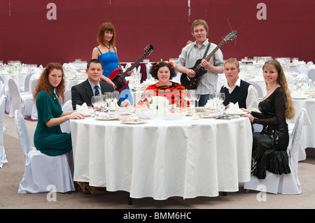 Fünf junge Leute am runden weißen Tisch im Restaurant und zwei Künstler mit Gitarren. Dinner-party Stockfoto