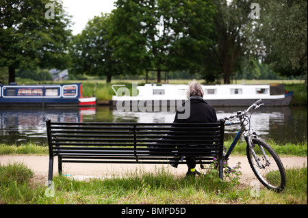 Eine Frau liest draußen auf einer Bank durch den Nocken in Cambridge mit ihrem Fahrrad neben ihr und schmale Boote im Hintergrund. Stockfoto