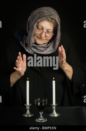 Eine ältere jüdische Frau, Kopf mit einem Tuch bedeckt, betet über zwei Sabbat-Kerzen und Sabbat Wein Stockfoto