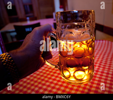 Nahaufnahme von einem Liter-Glas Lagerbier mit einem schaumigen Kopf in die Hand eines Mannes gegen eine rot-weiße Tischdecke und von hinten beleuchtet durch eine Kerze Stockfoto