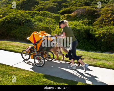 Eine gesunde junge Mutter von Zwillingen läuft auf einem steilen Hügel in Laguna Niguel, Kalifornien, drücken ihre Kinderwagen und zwei Hunde halten. Stockfoto