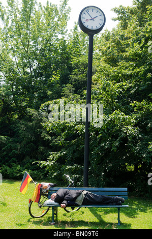 Mann im Anzug schläft auf Parkbank unter Uhr, neben ihm sein Fahrrad mit zwei deutschen Flaggen in Düsseldorf, Deutschland Stockfoto