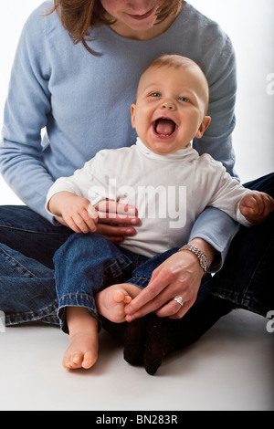 Kaukasische Babyjungen sitzt in seiner Mutter Schoß. Er lacht, weil sie seinen Fuß kitzeln ist. Stockfoto