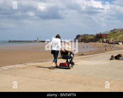 Eine junge Frau geht neben einem sehr alten Mann auf einem Elektromobil in Whitby North Yorkshire Stockfoto