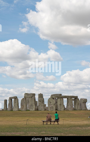 Mann und Frau sitzen auf einer Bank, Blick auf das berühmte Stonehenge prähistorische Monument an einem sonnigen Tag in Wiltshire, England Stockfoto