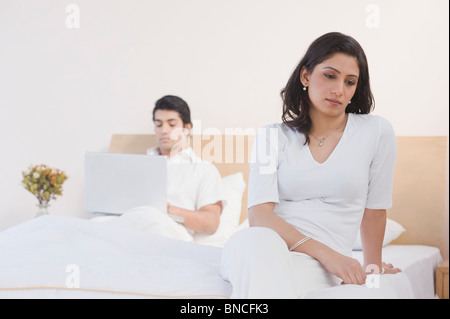 Ernsthafte Frau sitzt auf dem Bett und ein Mann, der auf einem Laptop arbeitet Stockfoto