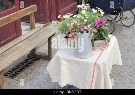 Ein einfaches und charmantes Arrangement aus frischen Wildblumen in einem Metallkrug sitzt auf einem Tisch mit Leinenbezug und verleiht einer französischen Straße einen rustikalen Touch. Stockfoto