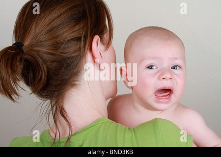 Weinen und beunruhigten jungen gehalten und getröstet von seiner Mutter vor einem einfarbigen Hintergrund. Das Baby ist Stockfoto