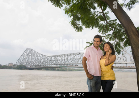 Paar steht am Fluss mit einer Brücke im Hintergrund, Howrah Bridge, Hooghly River, Kolkata, Westbengalen, Indien Stockfoto