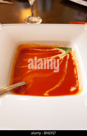 Eine samtige Tomatensuppe in einer quadratischen weißen Schüssel, garniert mit einem frischen Basilikumblatt und einem zarten Schuss Sahne Stockfoto