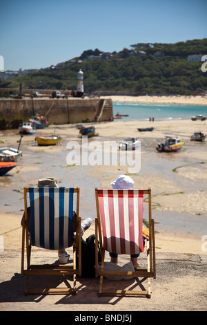 Altes Ehepaar mit Hüte auf sprechen Sie über die guten alten Zeiten während setzte sich auf den Liegestühlen am Hafen in St. Ives, Cornwall, England Stockfoto