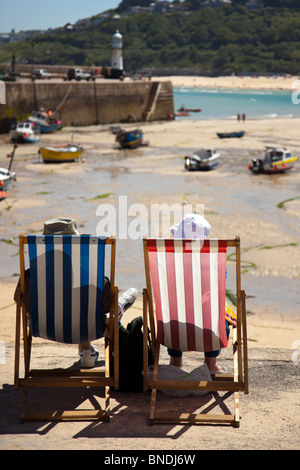 Altes Ehepaar mit Hüte auf sprechen Sie über die guten alten Zeiten während setzte sich auf den Liegestühlen am Hafen in St. Ives, Cornwall, England Stockfoto