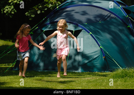 Zwei Mädchen laufen und Lachen in die Kamera mit einem großen Familienzelt im Hintergrund auf einem sonnigen Campingplatz. Stockfoto
