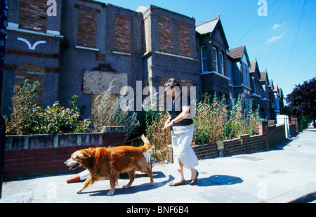 Als eine der wenigen Bewohner, die auf der Straße zurückgelassen wurden, geht eine Frau mit ihrem Hund an verroteten Häusern vorbei, die für die Sanierung gebaut wurden, Plumstead, South East London, Großbritannien Stockfoto