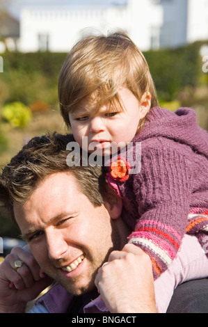 Vertikale Porträt eines jungen Vaters tragen seine süße Tochter auf den Schultern in der Sonne. Stockfoto