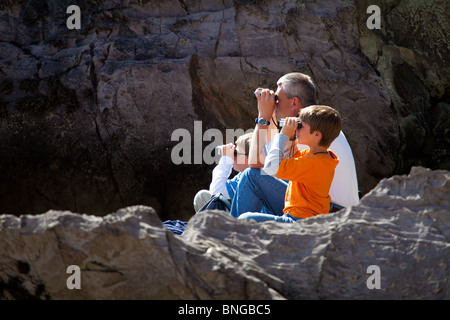 Ein Vater und zwei jungen sitzen auf Felsen und alle Blick auf das Meer mit dem Fernglas Stockfoto