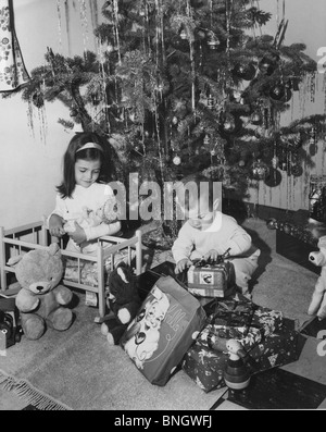 Mädchen spielen mit einer Puppe und ihr Bruder öffnen ein Weihnachtsgeschenk neben ihr, 1960er Jahre Stockfoto