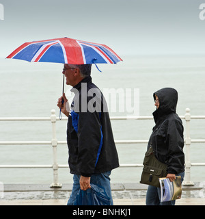 Menschen zu Fuß entlang Aberystwyth promenade im Regen eine Juli nass Sommernachmittag, Wales UK Union Jack Dach festhalten Stockfoto