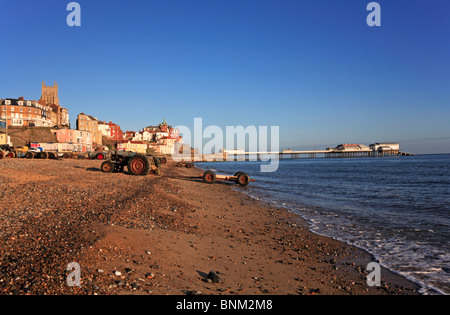 Fishermans Zugfahrzeuge und Anhänger an der Küste am Weststrand bei Cromer, Norfolk, England, UK, mit Pier im Hintergrund. Stockfoto