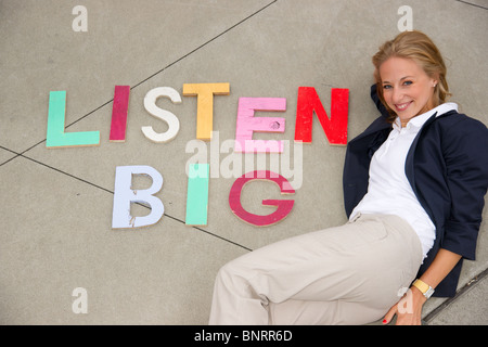 Attraktive blonde Frau posiert neben den Satz "Big hören" ausgeschrieben in Druckbuchstaben auf dem Boden neben ihr Stockfoto