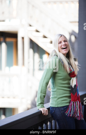 Eine junge Frau mit blonden Haaren lacht und lächelt beim Entspannen in der Nähe von einem Geländer an eine Skihütte. Stockfoto