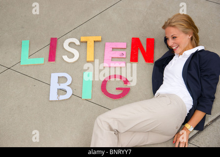 Attraktive blonde Frau posiert neben den Satz "Big hören" ausgeschrieben in Druckbuchstaben auf dem Boden neben ihr Stockfoto