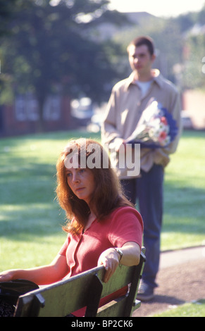 Frau sitzt auf einer Bank im Park im Vordergrund mit einem Mann mit einen Blumenstrauß zu ihr kommen Stockfoto