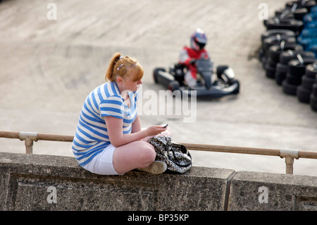 Junge Mädchen sass an der Wand mithilfe von Mobiltelefon während go kart Geschwindigkeiten durch im Hintergrund Stockfoto