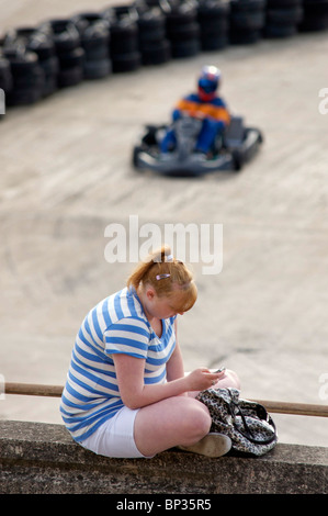 Junge Mädchen sass an der Wand mithilfe von Mobiltelefon während go kart Geschwindigkeiten durch im Hintergrund Stockfoto