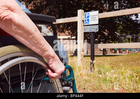 Ältere Frau im Rollstuhl durch ein Auto in behinderten Parkplatz durch ein Zeichen für blue badge Rollstuhlfahrer geparkt nur sitzen. England, Großbritannien, Großbritannien. Stockfoto