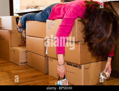 Eine erschöpfte Frau bricht zusammen auf einen Haufen von Umzugskartons nach Verpackung ganztägig.  Sie hält eine Kaffeetasse und einer Rolle Klebeband. Stockfoto