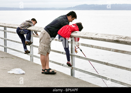 glücklicher asiatischen Vater zeigt kleine Tochter Umgangs mit Stab auf Edmonds Angelpier unter grauem Himmel, während jungen Sohn Uhren Stockfoto