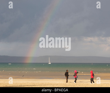 Menschen am Strand mit einem Regenbogen in der Ferne in Weymouth, Dorset, UK. Stockfoto