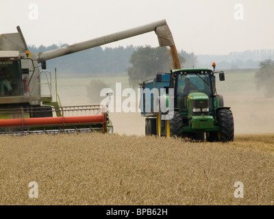 CLAAS Mähdrescher Harvester entladen Korn in einem Anhänger abgeschleppt von einem John Deere Traktor auf einem leicht bewölkten Sommertag Stockfoto